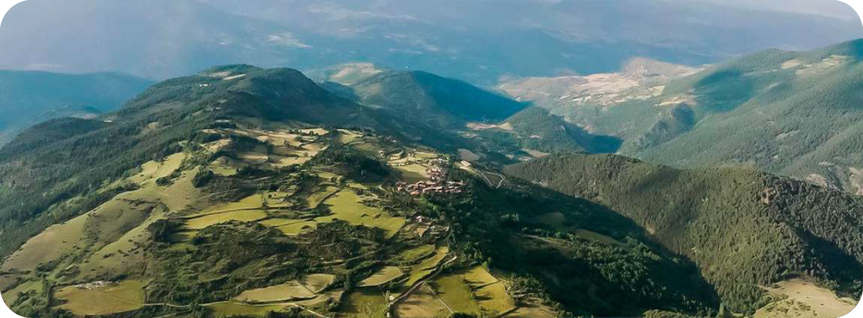 Luchtfoto van het bergdorp Montellà in de Spaanse Pyreneeën, omringd door groene landbouwvelden en beboste bergen onder een heldere lucht.