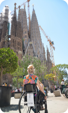 Een persoon in een rolstoel kijkt van een afstand omhoog naar de torens van de Sagrada Família in Barcelona onder een blauwe hemel.