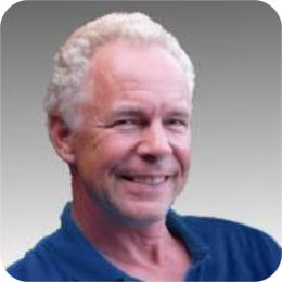 Portret van een oudere man met grijs haar, gekleed in een blauw overhemd, die vriendelijk in de camera lacht voor een zachte, onscherpe achtergrond.