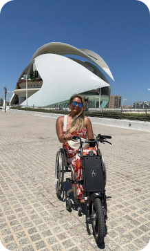 Een persoon in een rolstoel met een elektrische aankoppelfiets bevindt zich op het plein voor het futuristische, witte operagebouw Palau de les Arts Reina Sofía in Valencia onder een blauwe lucht.