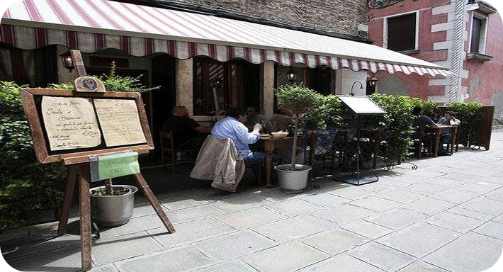 Het terras van restaurant Trattoria Storica in Venetië met een rood-wit gestreepte luifel, tafels buiten en een houten menukaartbord op de voorgrond.