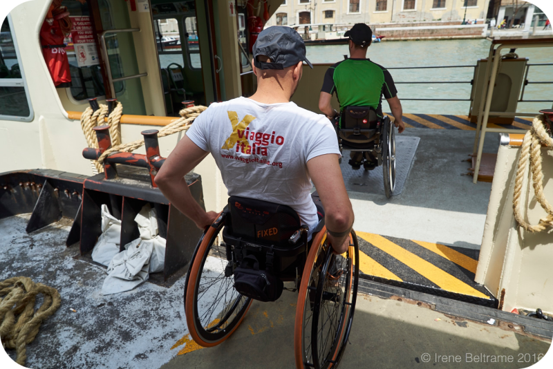 Twee mannen in rolstoelen gaan via een oprijplaat naar binnen in een  openbare waterbus in Venetië; één draagt een T-shirt van Viaggio Italia