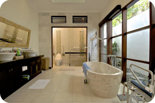 Ruime rolstoeltoegankelijke badkamer in Villa G at Ellora in Bali met vrijstaand stenen bad, plafondtillift, aangepast toilet met beugels en een doucheruimte met verzorgingstafel.