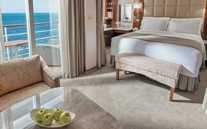 Luxe cruiseschip-suite met groot bed, zithoek, eettafel en balkon. Elegant ingericht met warme tinten en veel natuurlijk licht.