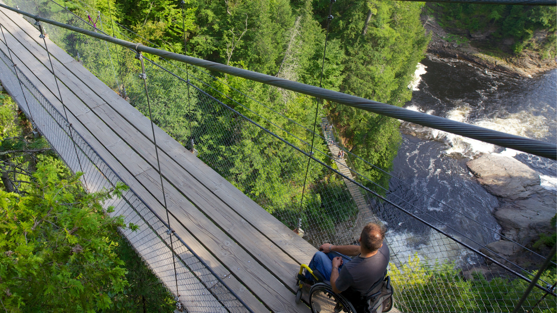 Man in rolstoel op houten hangbrug boven rivier in bosrijk gebied, laat toegankelijke natuurbeleving en avontuur in Canada zien.