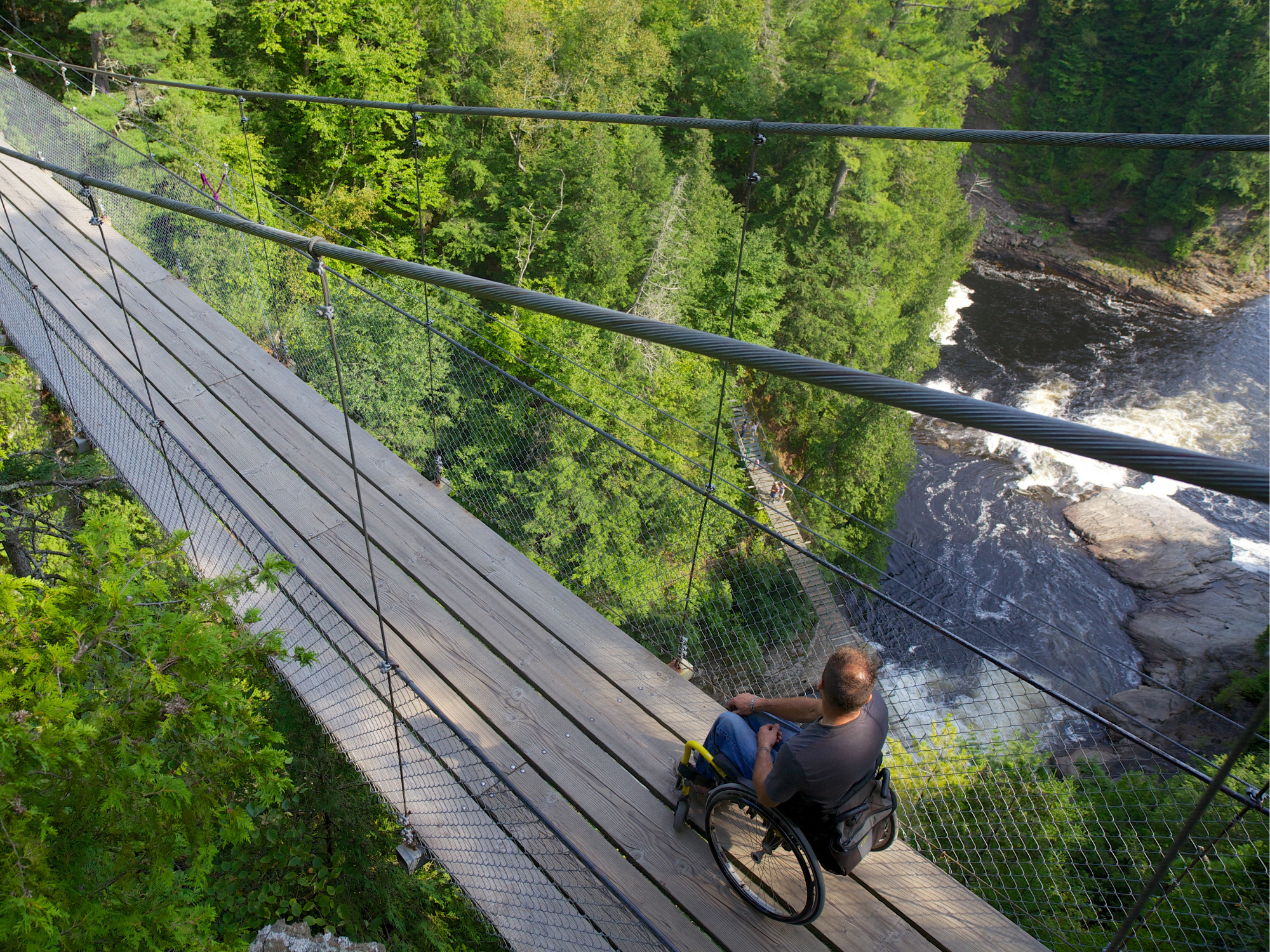 Man in rolstoel op houten hangbrug boven rivier in bosrijk gebied, laat toegankelijke natuurbeleving en avontuur in Canada zien.