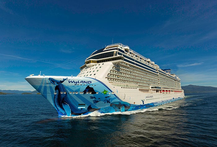 Cruiseschip Norwegian Bliss vaart over een kalme zee onder een heldere blauwe lucht, gezien vanaf de zijkant.