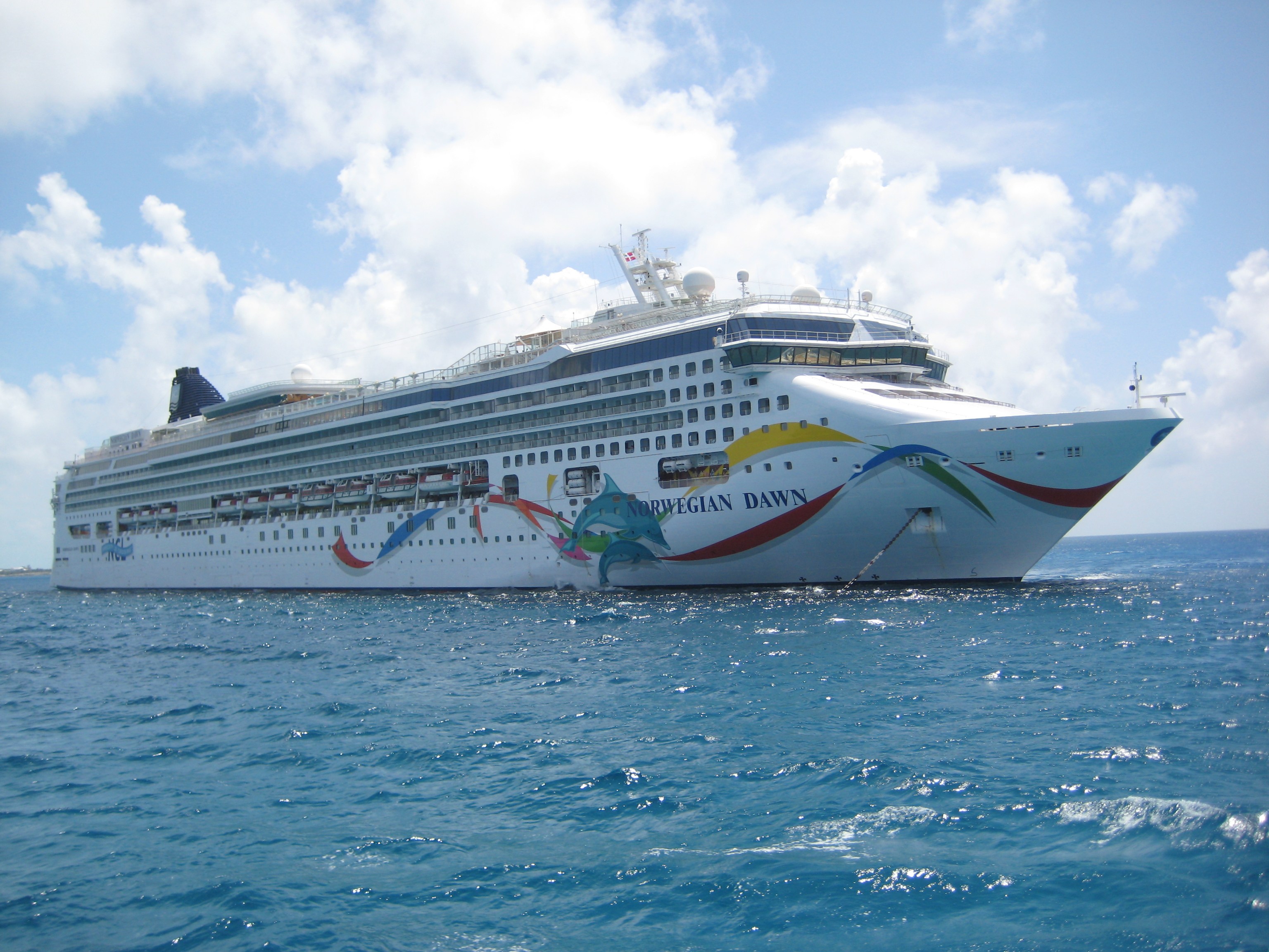 Cruiseschip Norwegian Dawn vaart over een kalme zee onder een heldere blauwe lucht, gezien vanaf de zijkant.