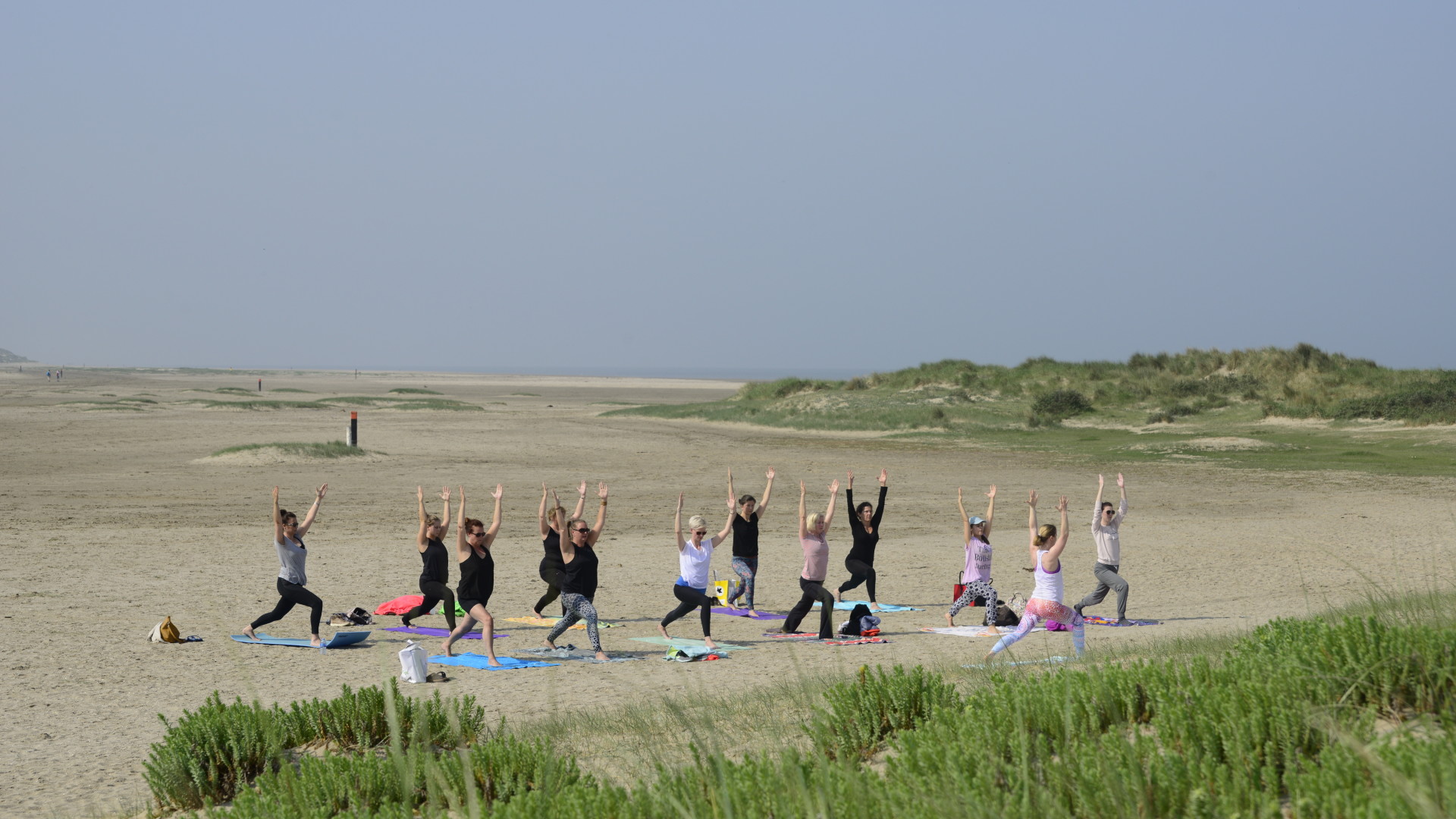 Groep yoga op Oostvoorne strand in yogahouding  krijger 1 - heldenhouding I - virabhadrasana I