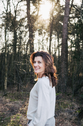 Zakelijk portret van Patricia, lachend over haar schouder terwijl ze staat in een bosrijke omgeving met tegenlicht van de ondergaande zon. Gefotografeerd door Fotografie Debbie