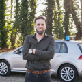 Gert-Jan Heerink van zijn rijschool, zelfverzekerd poserend met gekruiste armen voor zijn lesauto met lesauto-bord op het dak, omgeven door groen in tegenlicht. Zakelijke portretfotografie door Fotografie Debbie.