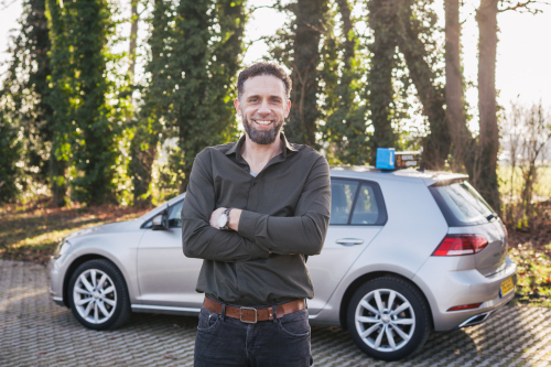 Gert-Jan Heerink van zijn rijschool, zelfverzekerd poserend met gekruiste armen voor zijn lesauto met lesauto-bord op het dak, omgeven door groen in tegenlicht. Zakelijke portretfotografie door Fotografie Debbie.