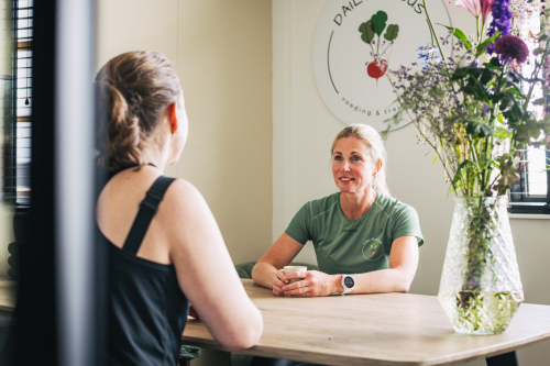 Sabine van Dailylicious voeding & training in gesprek met een klant aan een tafel, in haar eigen bedrijfsruimte met het Dailylicious-logo zichtbaar op de achtergrond. Authentieke zakelijke fotografie door Fotografie Debbie.
