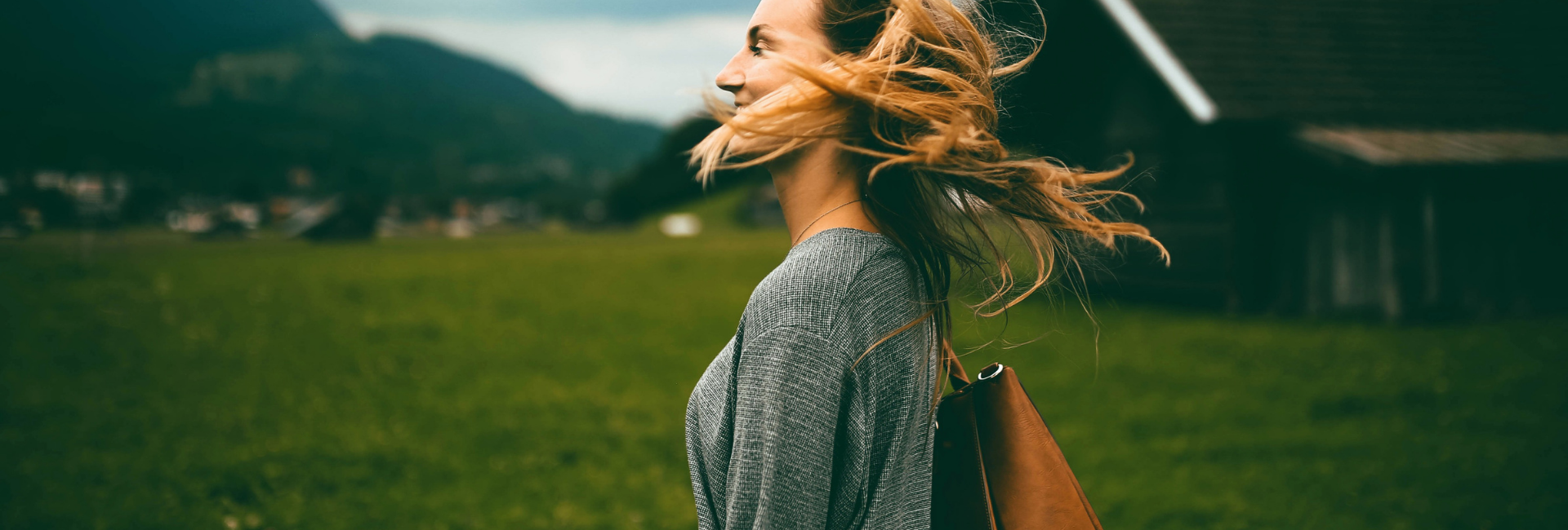 13 weken training lifestyle planning Vrouw met wapperend blond haar loopt ontspannen door een groen veld met op de achtergrond houten huisjes en bergen, symbool voor vrijheid, rust en balans.