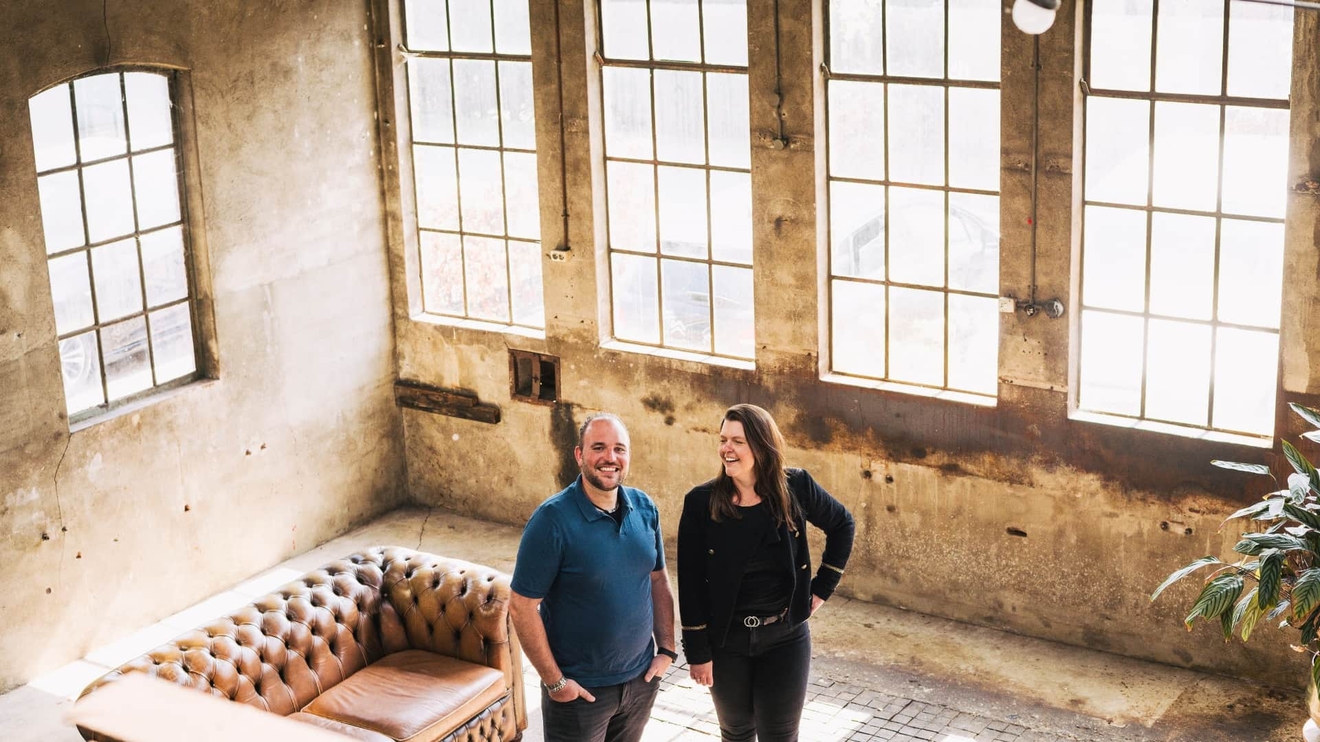 Harold en Lisette van Grenzeloos Gastvrij lachen samen in een industriële ruimte, symbool voor hun persoonlijke en nuchtere aanpak voor horecaondernmers