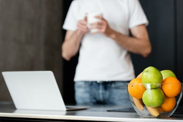 Man met een koffie mok die staat achter een laptop en fruitschaal