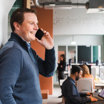 Dennis lachend aan de telefoon. Collega's aan het werk op de achtergrond.