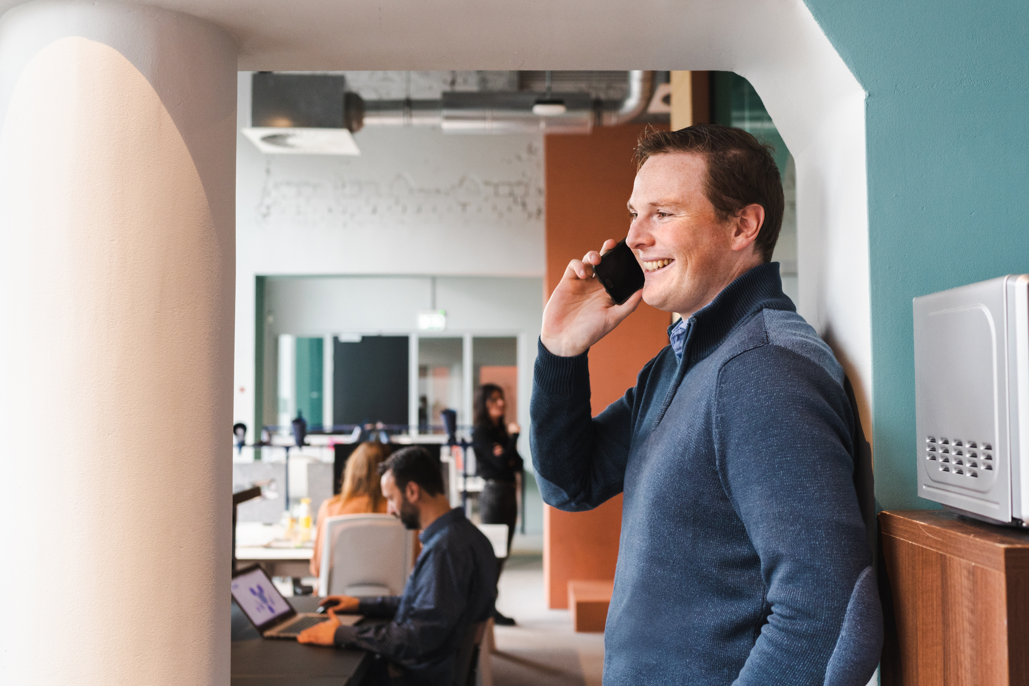Dennis lachend aan de telefoon. Collega's aan het werk op de achtergrond.