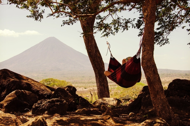 Persoon ligt in een rode hangmat in de schaduw, gespannen tussen twee bomen en een berg op de achtergrond