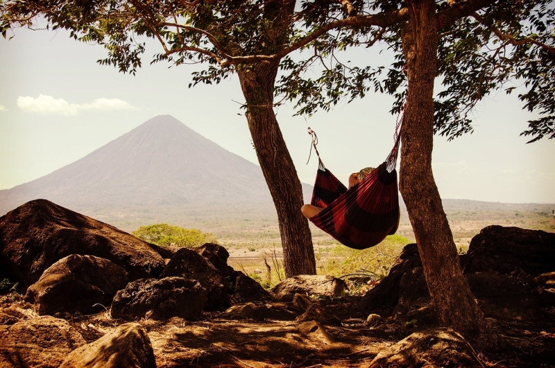 Persoon ligt in een rode hangmat in de schaduw, gespannen tussen twee bomen en een berg op de achtergrond