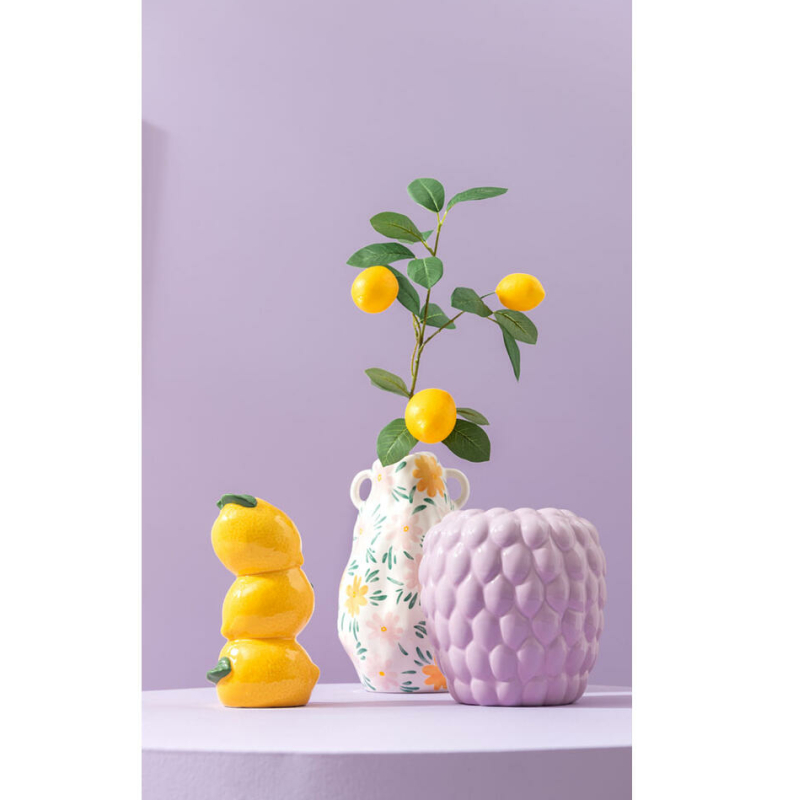 een klein geel citroenvaasje staat op een lila tafel samen met twee andere vazen met een citroentak