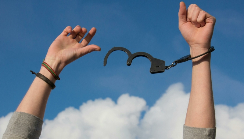 Je ziet twee armen en handen in de lucht, waarbij 1 hand uit de handboeien is losgekomen omdat de handboei openstaat en de ander nog in de handboei zit.