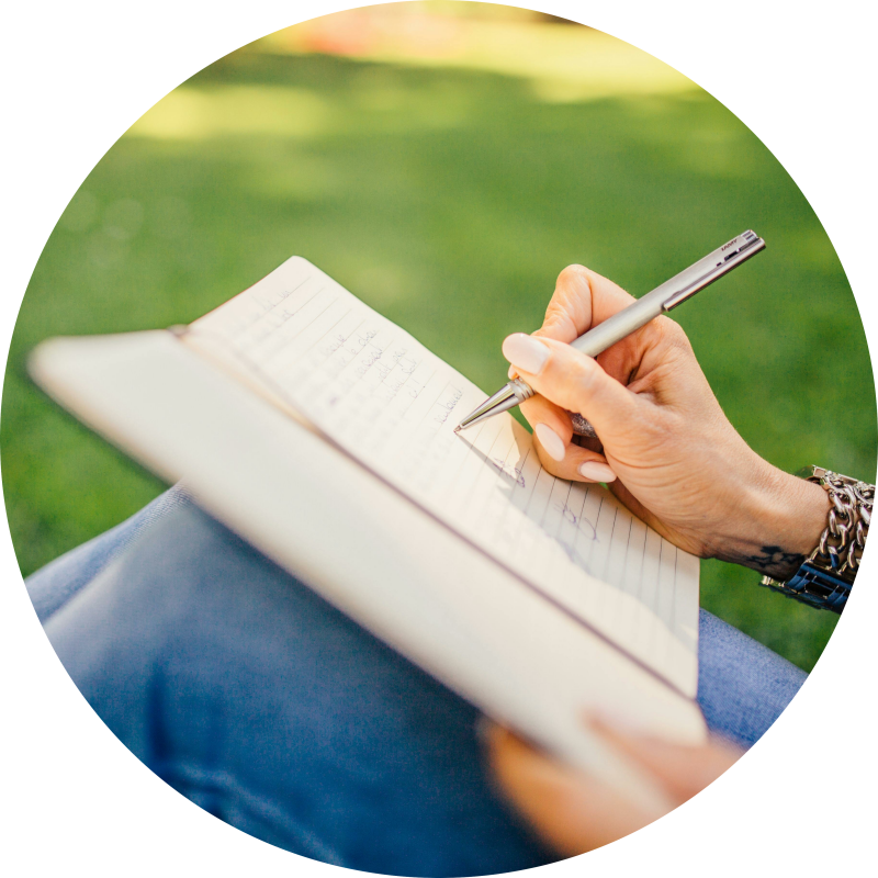 Je ziet een vrouw op het gras zitten met een journal op haar knieen waar ze in schrijft.