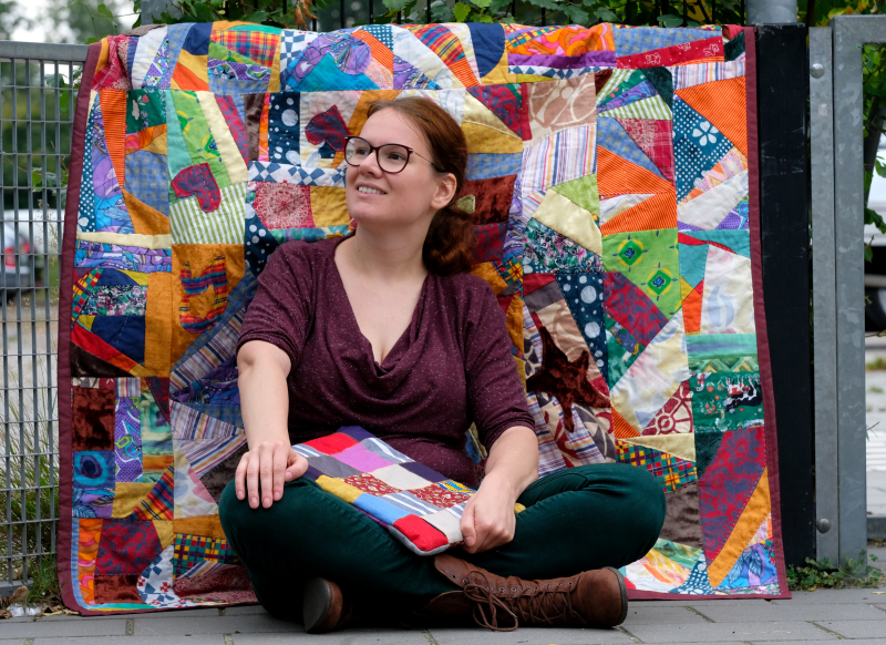 Quilter Rianne sitting croslegged with a colourful crazy quilt as backdrop and a quilted laptop sleeve on her lap