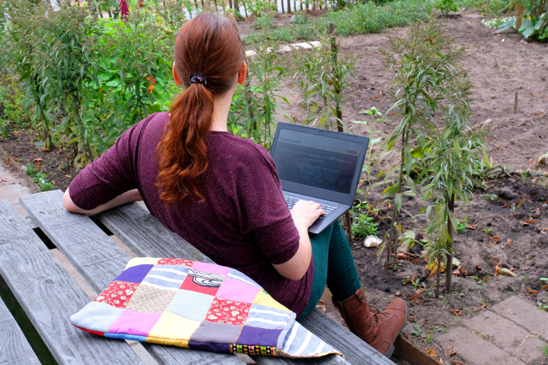 Rianne working in garden on laptop with red hair and red shirt. On the bench is a laptop sleeve