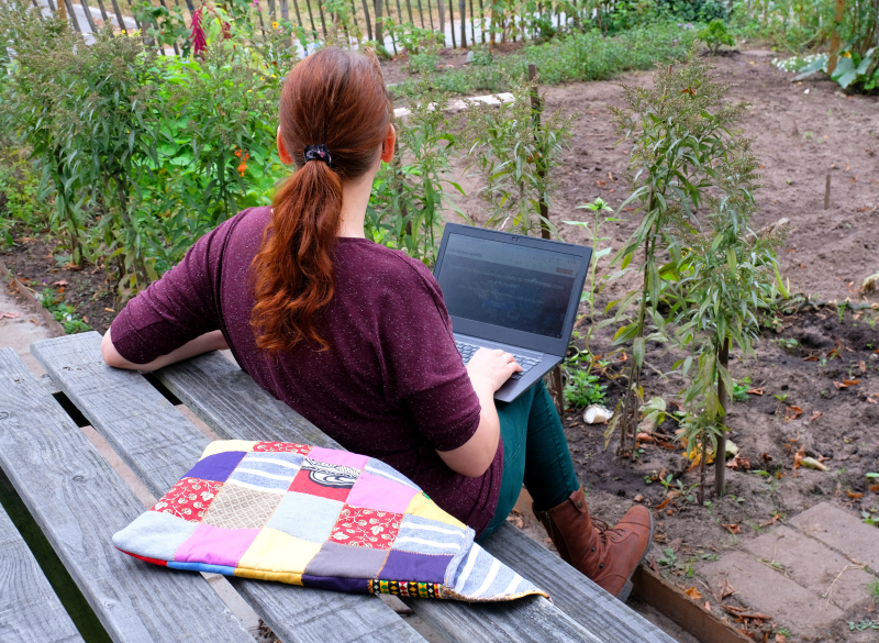 Rianne working in garden on laptop with red hair and red shirt. On the bench is a laptop sleeve