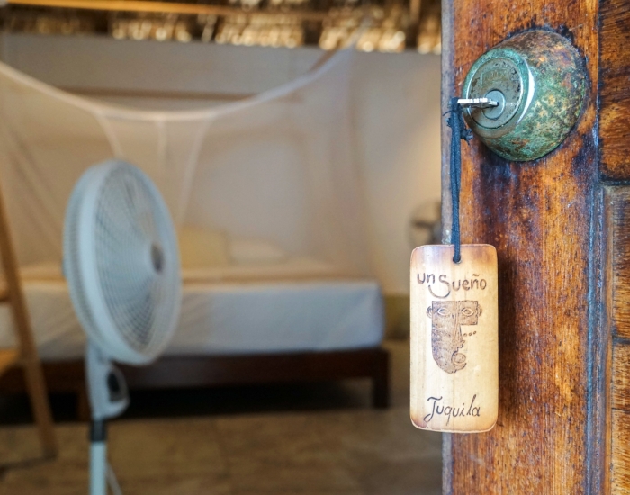 Close-up van houten deur met Un Sueño sleutelhanger van cabin Juquila, met ventilator en bed met klamboe zichtbaar op de achtergrond