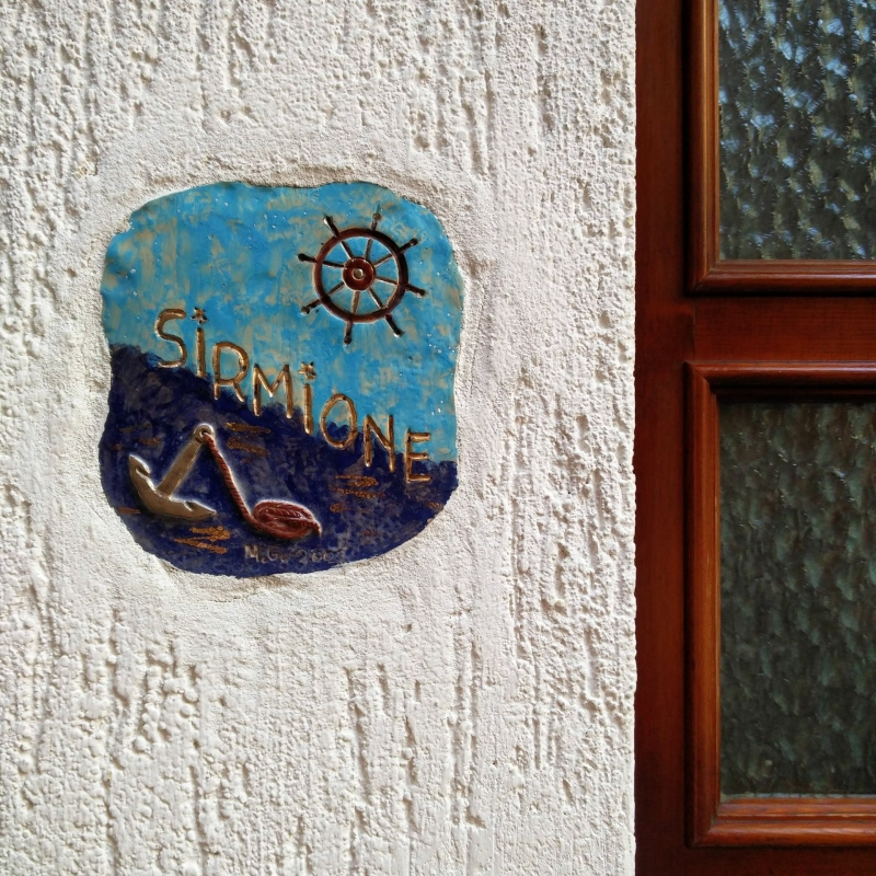 Blauw keramiek tegeltje met “Sirmione”, anker en stuurwiel op een witte muur