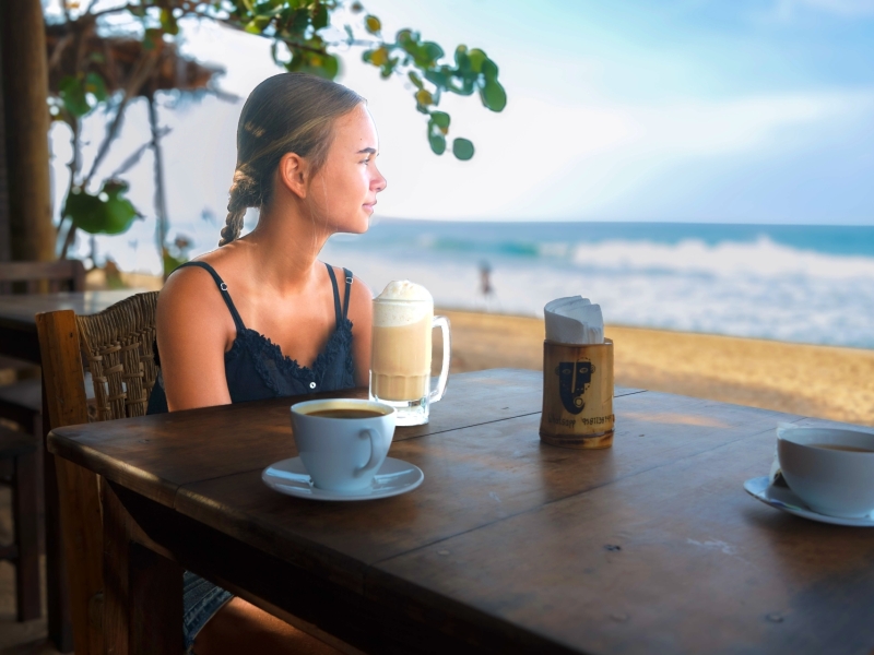 Meisje drinkt koffie aan een houten tafel aan het strand bij restaurant Un Sueño, met de golven van de Stille Oceaan op de achtergrond
