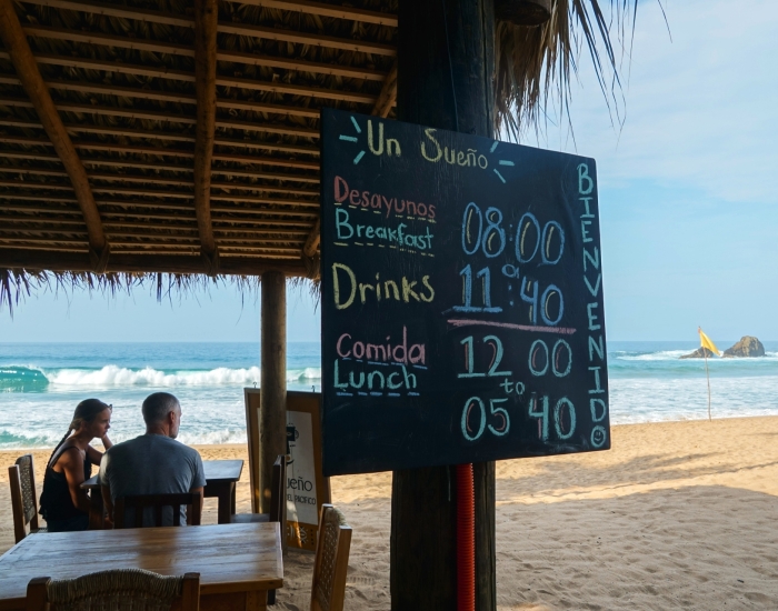 Krijtbord met openingstijden van strandrestaurant Un Sueño in San Agustinillo, met koppel en golven van de Stille Oceaan op de achtergrond