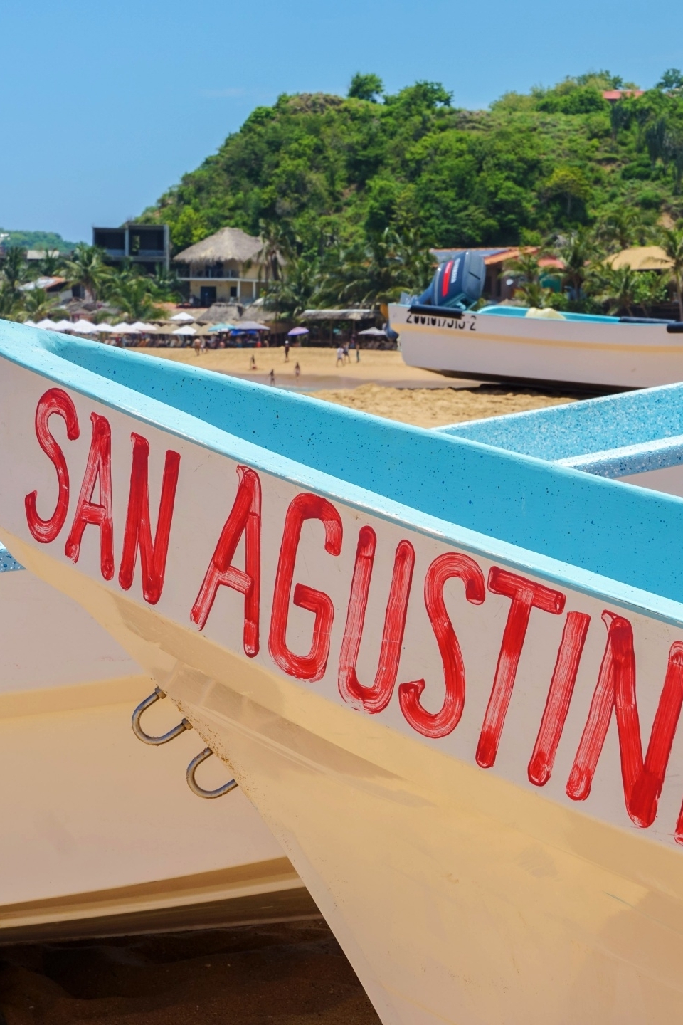 Kleurrijke vissersbootjes met de naam San Agustinillo op het strand, met palmbomen en kustlijn op de achtergrond