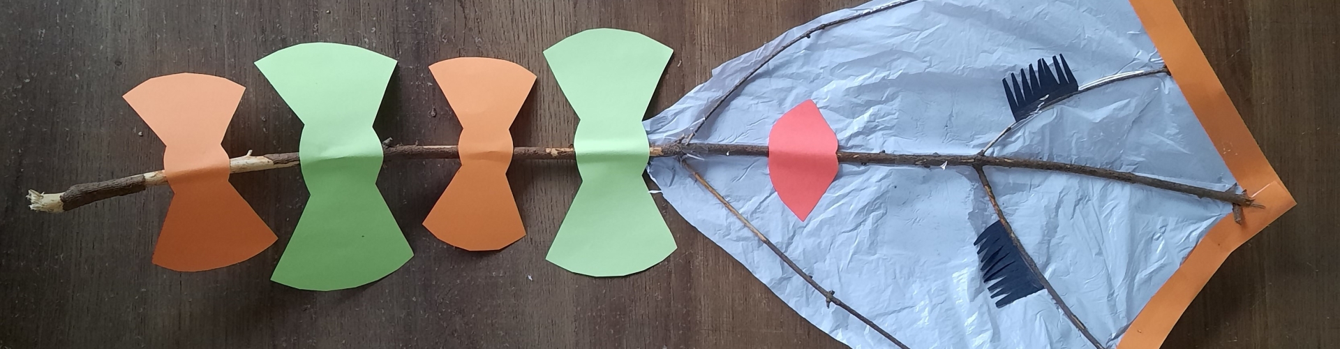 Een decoratieve vlieger gemaakt van takken en oud plastic en restjes knutselpapier waarvan ogen, een mond en strikjes zijn gemaakt