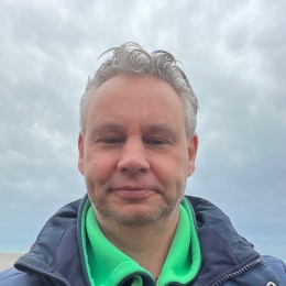 Portretfoto van een rustige, vriendelijke man buiten in de natuur. Hij heeft een zacht, ontspannen gezicht en draagt een donkerblauwe jas met een groene trui. Achter hem is een open lucht zichtbaar met wolken, wat een gevoel van ruimte en eenvoud geeft.