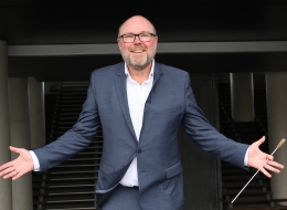 Alexander de Blaeij in blauw pak met open armen en een dirigeerstok in de hand, lachend voor een trappenhuis. De foto straalt enthousiasme en uitnodiging uit.
