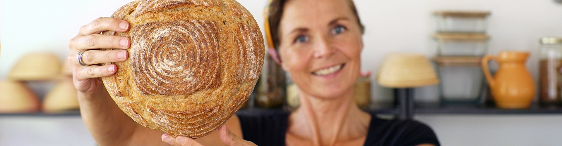 Zelf brood bakken - Marije Bakt Brood lacht met ambachtelijk brood voor zich uit gestoken in bakkers keuken met potjes