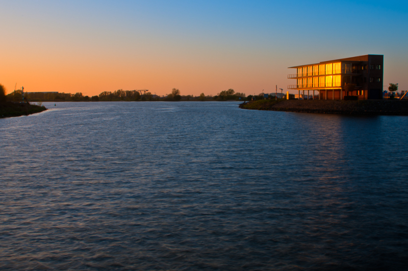 Warme zonsondergang bij een modern gebouw aan de oever van een brede rivier, met een kleurrijke lucht en zacht golvend water – een voorbeeld van landschapsfotografie met een lage horizon voor veel lucht en rust.