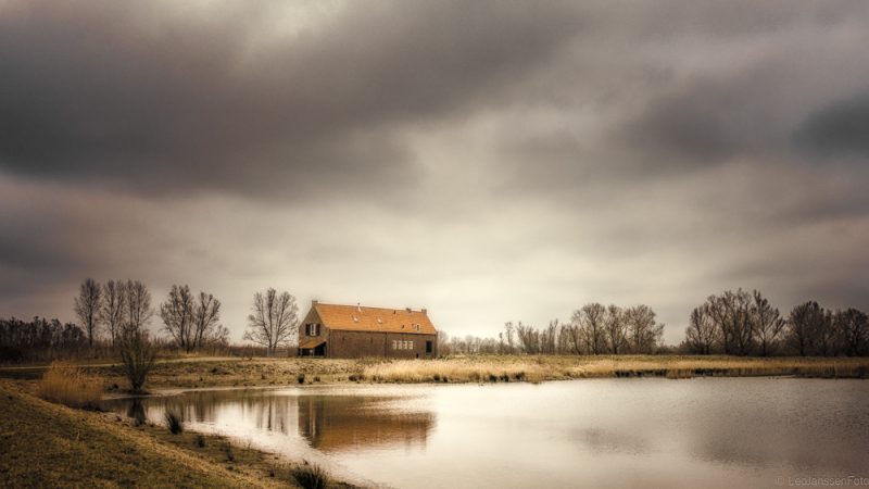 Rustiek landschap met een boerderij aan een plas water, waarin de lucht dramatisch weerspiegeld wordt. De horizon ligt hoog, waardoor de nadruk op het lichtspel in de lucht en reflectie komt te liggen.