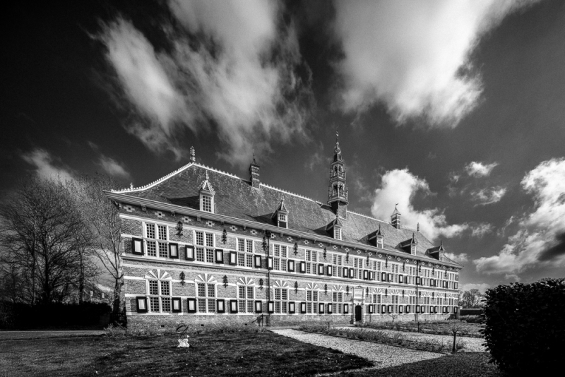 Een krachtig zwart-witbeeld van een symmetrisch, historisch pand onder een expressieve wolkenlucht. Het lage camerastandpunt en de strakke lijnen benadrukken het perspectief en geven de foto een grafisch karakter.