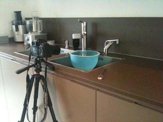 Camera op statief gericht op een druppelende kraan boven een blauwe schaal, als voorbeeld van een eenvoudige opstelling voor druppelfotografie