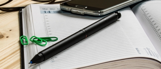 Een opengeslagen agenda waarop een pen, een telefoon en wat paperclips liggen.