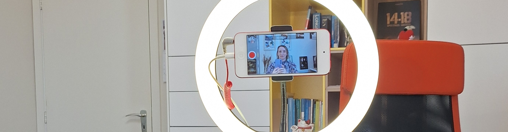 video-opnames voor de online audiodescriptie-workshop Een statief met ringlamp waarin met een camera in mijn thuiskantoor. Wat verder in de kamer staat een oranje bureaustoel met een poezenkussen en een hoge, witte boekenkast met gele en houtkleurige accenten.