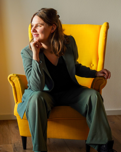 Nienke Vink zittend in een gele stoel, kijkend naar buiten, met een glimlach en een professionele uitstraling. Dit portret weerspiegelt haar expertise in carnivoor diëten en dierlijke voeding.