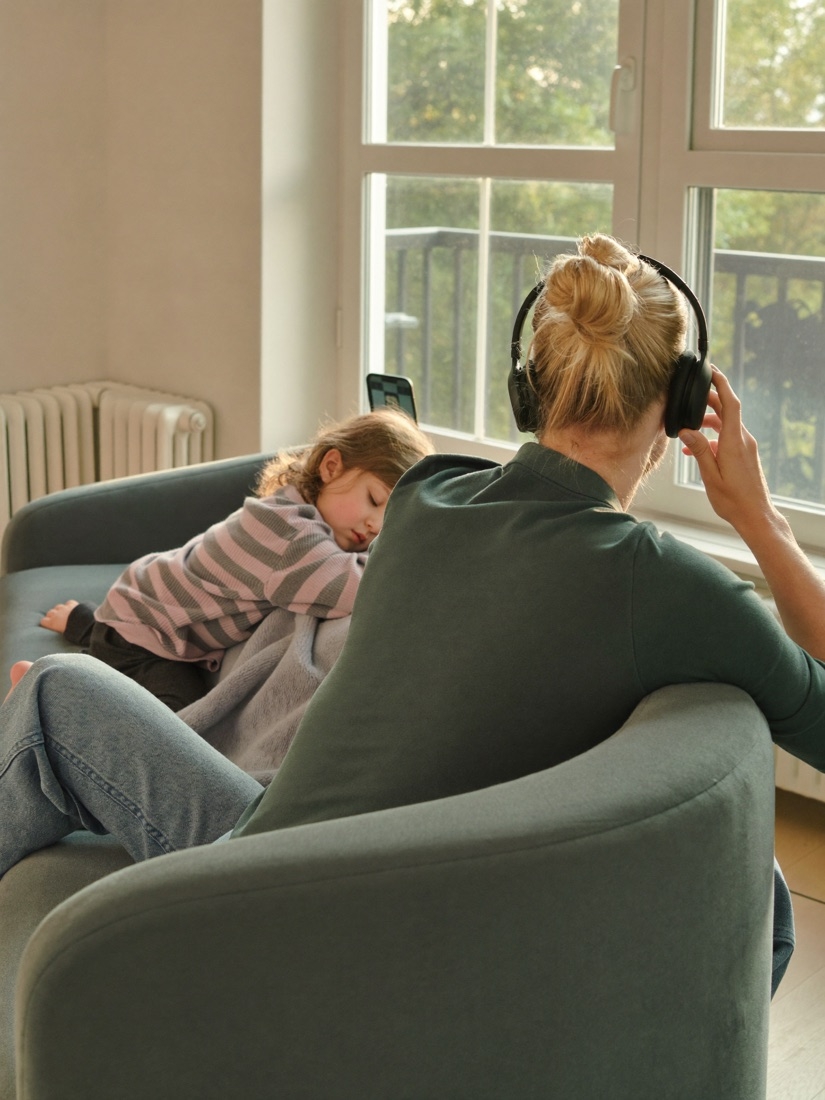 Ouder op de bank met een koptelefoon op, een kind slaapt tegen haar aan