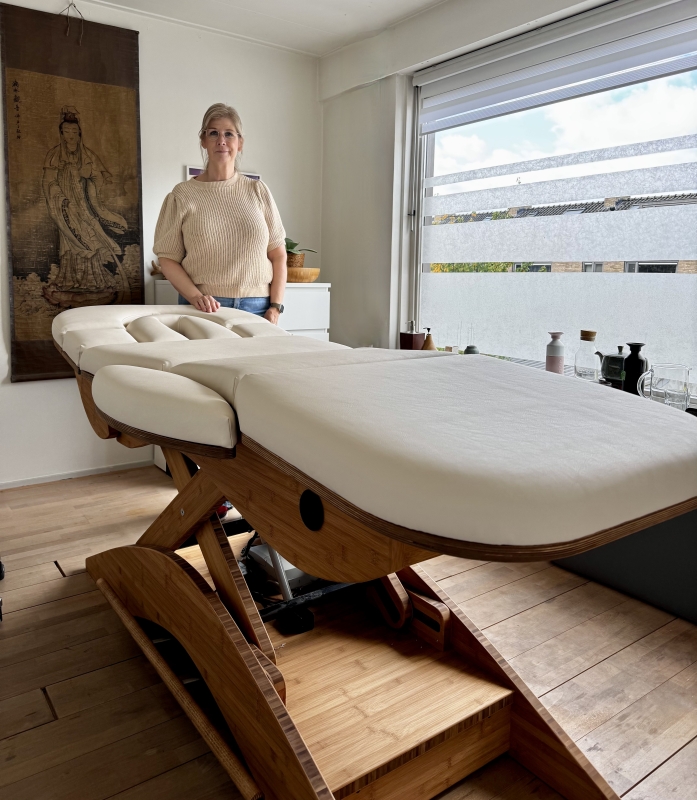 Massageruimte met houten elektrische massagetafel en therapeut, natuurlijk licht en rustgevende inrichting.