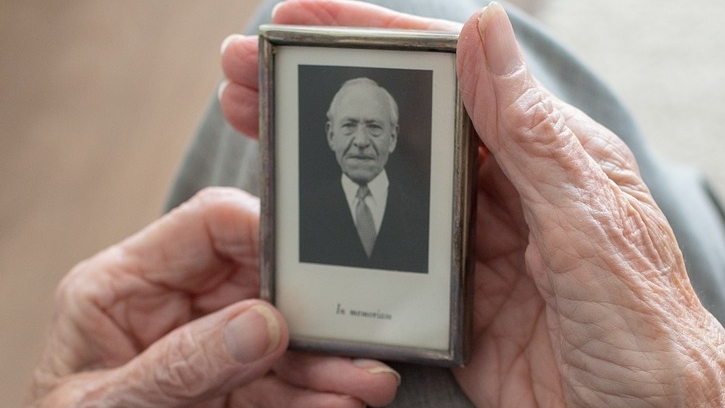Oudere vrouw met foto van haar man op schoot, in haar vertrouwde huis