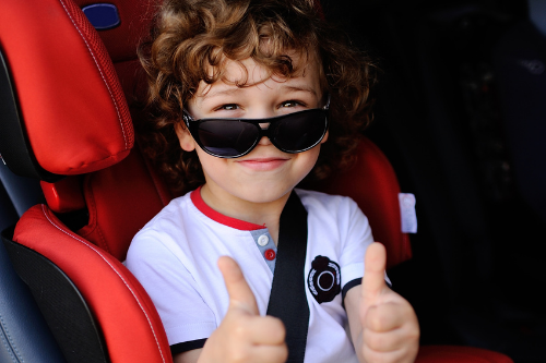 Jongen in de wagen in rode kinderstoel met zonnebril en steekt zijn duimpjes omhoog